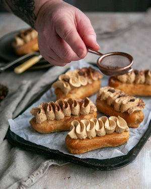 Honeywell Bakes Coffee Eclair Baking Kit - Person dusting chocolate powder over coffee eclairs on a tray