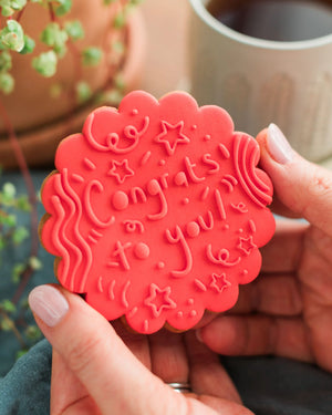 Honeywell Bakes Congrats To You Biscuit - Red cookie with 'Congratulations' text held by a hand, with a blurred background of a plant and mug.