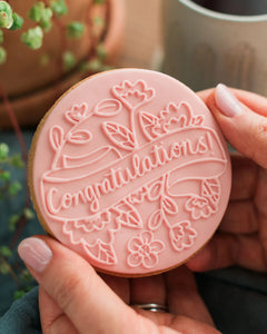 Honeywell Bakes Congratulations Biscuit - Person holding a pink 'Congratulations' cookie with floral designs.