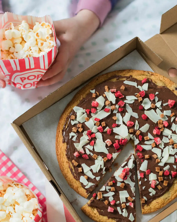 Honeywell Bakes Cookie Dough Pizza Baking Kit - Chocolate cookie pizza with toppings in a box, accompanied by popcorn, on a checkered tablecloth.