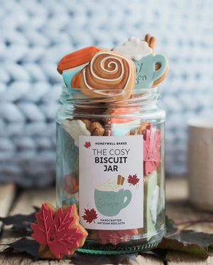 Honeywell Bakes The Cosy Biscuit Jar - A glass jar filled with assorted decorated cookies and surrounded by fall-themed props, labeled 'The Cosy Biscuit Jar' by Honeywell Bakes, set on a rustic wooden surface with a soft, blurred background.