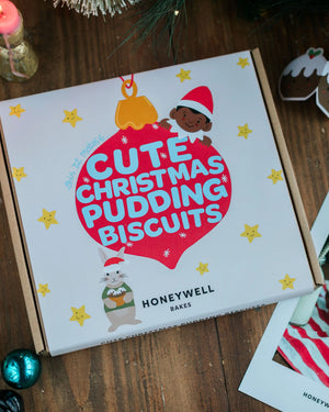 A Honeywell Bakes baking kit of Christmas-themed pudding biscuits, featuring festive holiday illustrations on a white lid, set on a rustic wooden table.