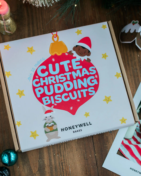 A Honeywell Bakes baking kit of Christmas-themed pudding biscuits, featuring festive holiday illustrations on a white lid, set on a rustic wooden table.