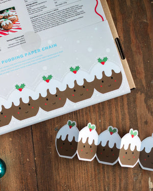 A holiday-themed paper chain made of pudding-shaped cutouts with white icing, brown filling, and holly accents, from Honeywell Bakes Cute Christmas Sandwich Biscuit baking kit on a wooden table next to an open recipe card.