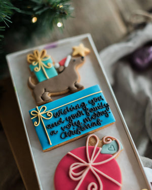 Honeywell Bakes Driving Home For Christmas Personalised Biscuit Gift - A close-up of decorated Christmas cookies arranged in a clear tray, including a dachshund with a gift box, a blue cookie with cursive holiday message, and a pink ornament cookie, with blurred pine branches and fabric in the background.