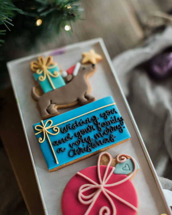 Honeywell Bakes Driving Home For Christmas Personalised Biscuit Gift - A close-up of decorated Christmas cookies arranged in a clear tray, including a dachshund with a gift box, a blue cookie with cursive holiday message, and a pink ornament cookie, with blurred pine branches and fabric in the background.