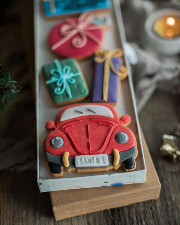 Honeywell Bakes Driving Home For Christmas Personalised Biscuit Gift - A long rectangular tray holds several decorated cookies shaped like gift boxes and a red Santa car, arranged on a wooden surface with a blurred candle and fabric in the background.