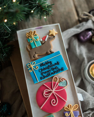 Honeywell Bakes Driving Home For Christmas Personalised Biscuit Gift - A tray of decorated Christmas cookies, including a dachshund with a gift box and star, a blue cookie with 'Merry Christmas from the Whittakers' in black script, a pink gift box with white bow details, and other holiday-themed cookies, styled on a rustic wooden table with a candle and greenery.