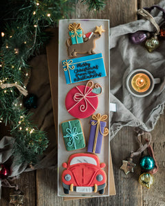 Honeywell Bakes Driving Home For Christmas Personalised Biscuit Gift - A rectangular tray of decorated Christmas cookies arranged vertically on a rustic wooden table, surrounded by holiday decor including evergreen branches, ornaments, a candle, and draped fabric.