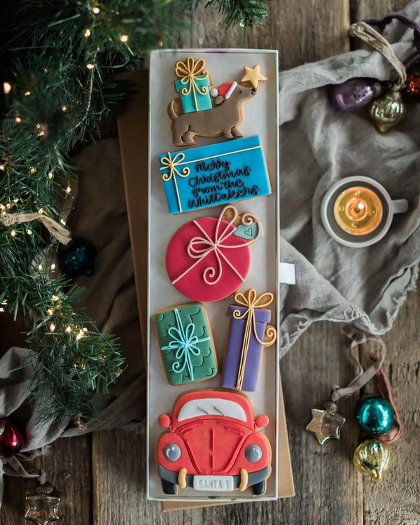 Honeywell Bakes Driving Home For Christmas Personalised Biscuit Gift - A rectangular tray of decorated Christmas cookies arranged vertically on a rustic wooden table, surrounded by holiday decor including evergreen branches, ornaments, a candle, and draped fabric.
