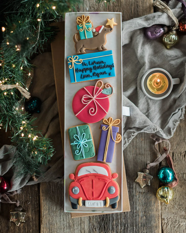 Honeywell Bakes Driving Home For Christmas Personalised Biscuit Gift - A long rectangular box of decorated Christmas-themed cookies arranged vertically, photographed from above on a rustic wooden surface with holiday decor including a candle, greenery, and ornaments around it.