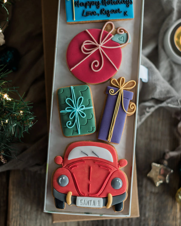 Honeywell Bakes Driving Home For Christmas Personalised Biscuit Gift - A boxed assortment of decorated sugar cookies shaped like holiday gifts and a red Santa car, arranged in a clear display tray on a rustic wooden surface with holiday decor around it.