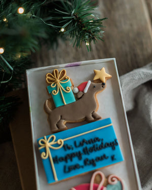 Honeywell Bakes Driving Home For Christmas Personalised Biscuit Gift - A close-up of a decorated holiday cookie shaped like a dachshund carrying a turquoise gift box with gold icing and a star, placed in a gift box beside a blue personalised message cookie and Christmas greenery with warm string lights.