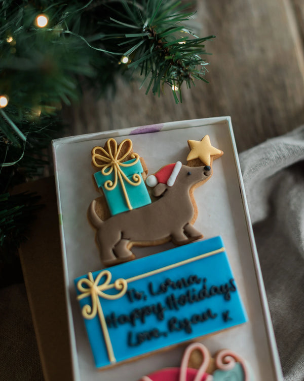 Honeywell Bakes Driving Home For Christmas Personalised Biscuit Gift - A close-up of a decorated holiday cookie shaped like a dachshund carrying a turquoise gift box with gold icing and a star, placed in a gift box beside a blue personalised message cookie and Christmas greenery with warm string lights.