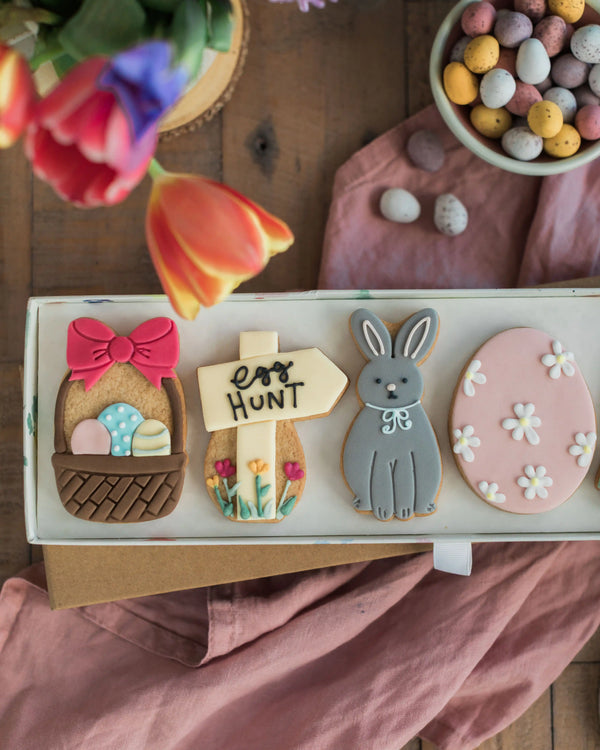 Honeywell Bakes Easter Egg Hunt Biscuit Gift - Decorative cookies shaped like a basket, sign, rabbit, and egg on a wooden surface with tulips and Easter eggs.