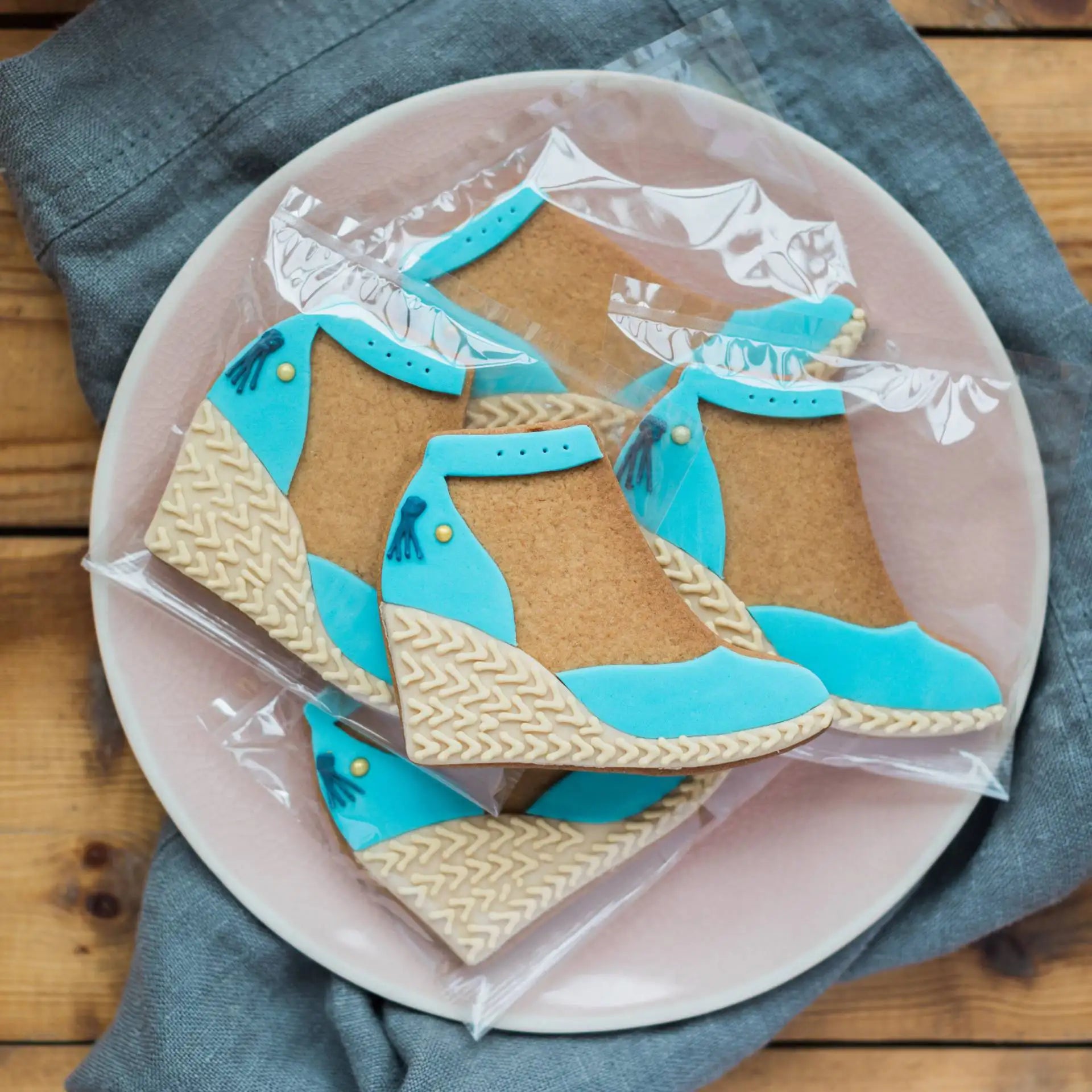Decorative custom cookies shaped like shoes with blue detail, made for Fairfax and Favour on a plate with a wooden background