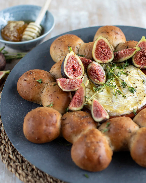Honeywell Bakes Fig and Walnut Bread Wreath Baking Kit - Baked camembert with figs and bread rolls on a dark plate with a bowl of honey and a stick in the background.