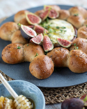 Honeywell Bakes Fig and Walnut Bread Wreath Baking Kit - Baked camembert with figs and bread rolls on a dark plate with a bowl of honey and a stick in the background.