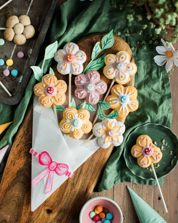 Honeywell Bakes Flower Biscuit Pops Baking Kit - Decorative cookies shaped like flowers on sticks arranged on a wooden board with a green cloth background.