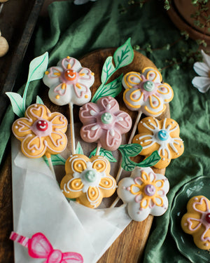 Honeywell Bakes Flower Biscuit Pops Baking Kit - Decorative cookies shaped like flowers on sticks arranged on a wooden board with a green cloth background.