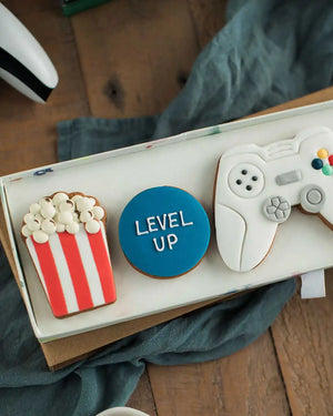 Honeywell Bakes Gamers Biscuit Gift Set - Decorative cookies shaped like a game controller, popcorn, and a blue circle with 'LEVEL UP' text on a wooden surface.