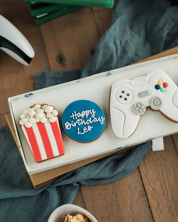 Honeywell Bakes Gamers Biscuit Gift Set - Three cookies shaped like a game controller, popcorn, and a blue circle with 'Happy Birthday Leo' text on a wooden surface.