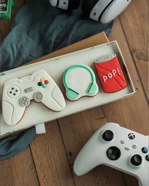Honeywell Bakes Gamers Biscuit Gift Set - Decorative cookies shaped like a gaming controller, headset, and can of pop on a wooden surface.