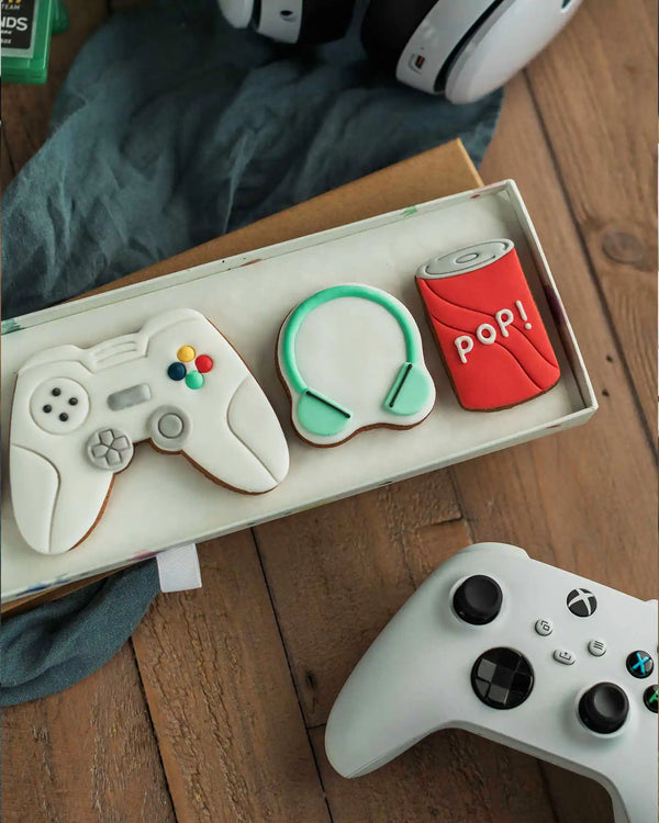 Honeywell Bakes Gamers Biscuit Gift Set - Decorative cookies shaped like a gaming controller, headset, and can of pop on a wooden surface.