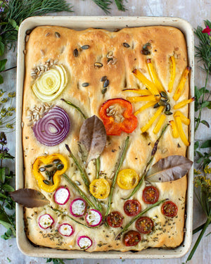 Honeywell Bakes Garden Focaccia Bread Baking Kit - Focaccia bread with various vegetables arranged on top, surrounded by greenery.