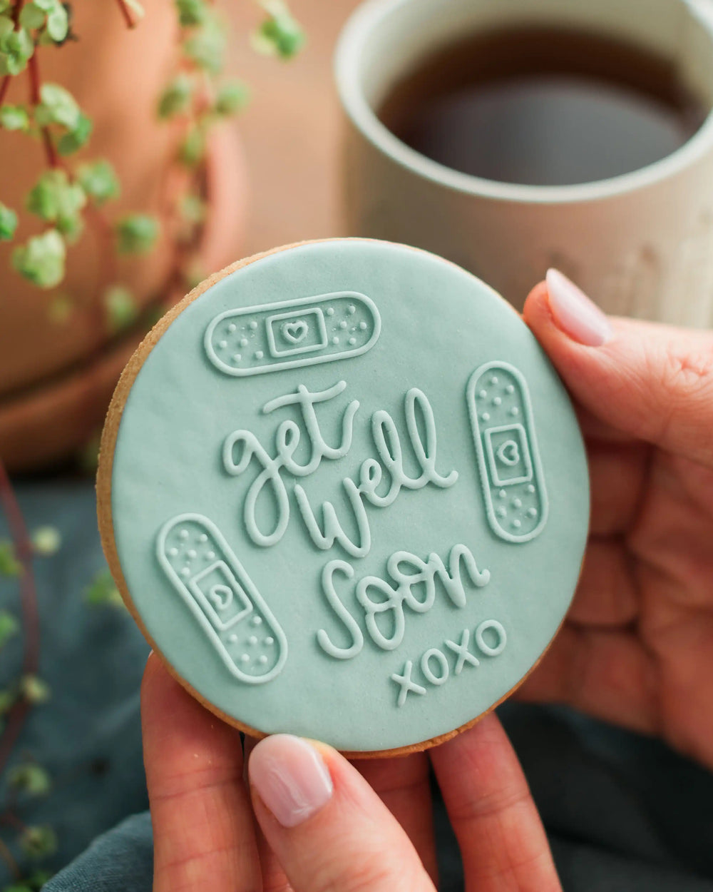 Honeywell Bakes Get Well Soon Biscuit - Cookie with 'get well soon' message held by a hand, with a cup of coffee and plant in the background.