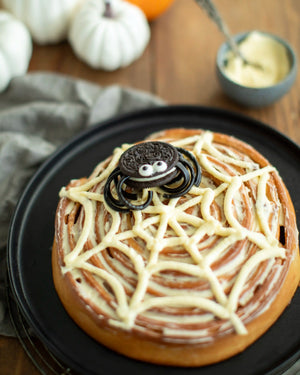 Honeywell Bakes - A Halloween-themed cinnamon roll decorated with white icing web patterns and topped with an Oreo cookie spider, served on a black plate.