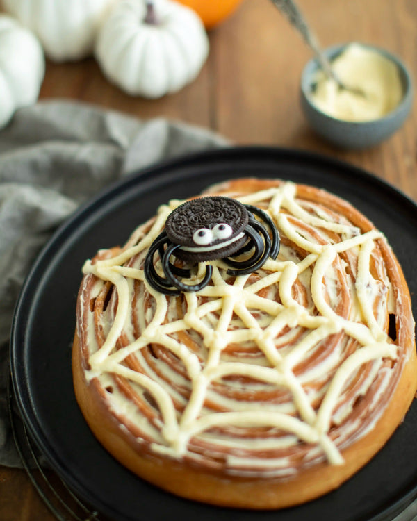 Honeywell Bakes - A Halloween-themed cinnamon roll decorated with white icing web patterns and topped with an Oreo cookie spider, served on a black plate.