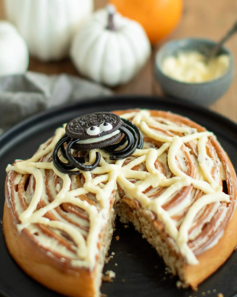 Honeywell Bakes - A Halloween-themed cinnamon roll cake on a black plate, decorated with white frosting in a web pattern and topped with a spider cookie. A slice is cut from the cake, and the background features pumpkins and a small bowl of frosting on a wooden table.