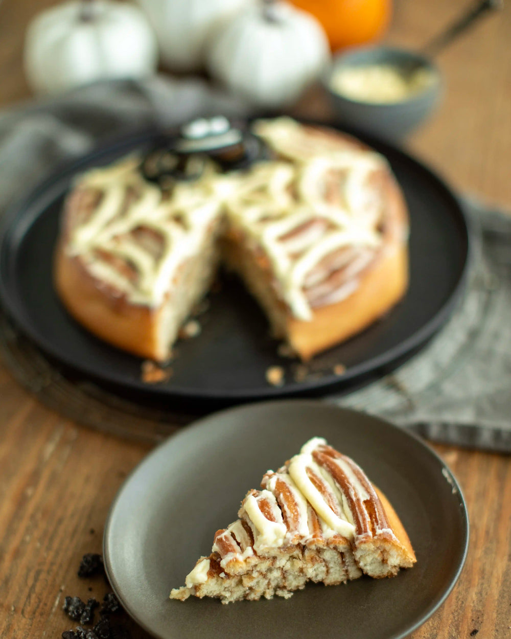Honeywell Bakes - A Halloween-themed cinnamon roll cake on a black plate, decorated with white frosting in a web pattern and topped with a spider cookie. A slice is cut from the cake, and the background features pumpkins and a small bowl of frosting on a wooden table.