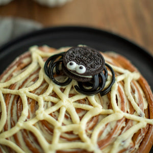 Honeywell Bakes - A close-up of a spider-themed cinnamon roll topped with white icing web patterns, a small Oreo cookie spider decoration with googly eyes, and black candy legs on a dark plate.