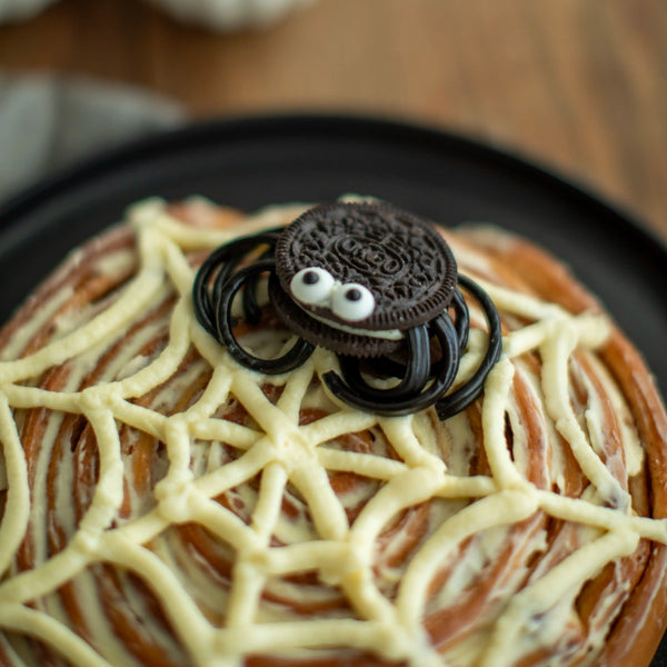 Honeywell Bakes - A close-up of a spider-themed cinnamon roll topped with white icing web patterns, a small Oreo cookie spider decoration with googly eyes, and black candy legs on a dark plate.