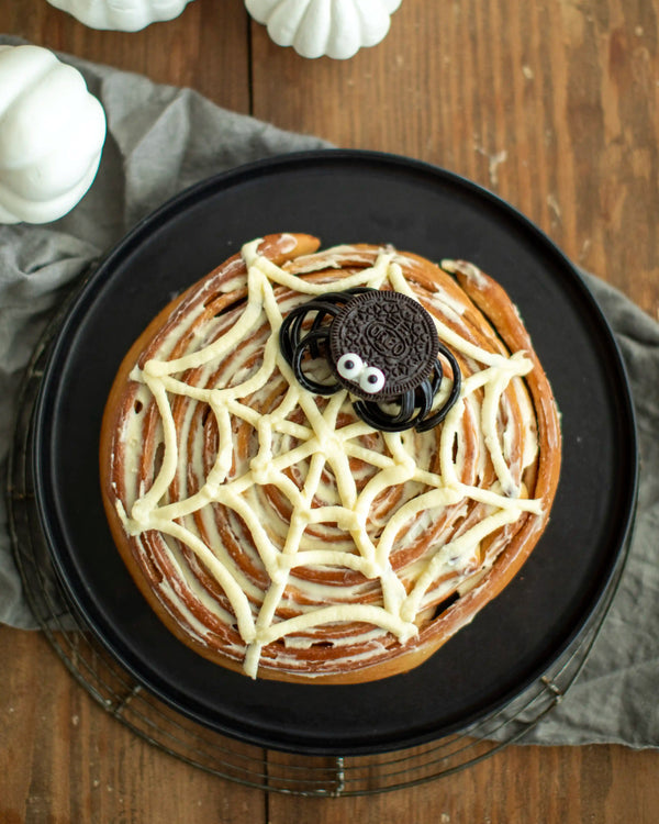 Honeywell Bakes - A Halloween-themed cinnamon roll decorated with white icing in a spider web pattern and topped with a chocolate Oreo cookie, presented on a black plate over a cooling rack on a wooden table.