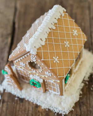 Honeywell Bakes Gingerbread House Baking Kit - A decorated gingerbread house with white icing snow, a white icing grid pattern, and green wreath accents, set on a rustic wooden surface with white shredded coconut base.