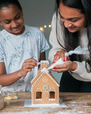 Honeywell Bakes Gingerbread House Baking Kit - A woman and a young girl decorating a gingerbread house together on a wooden table, with white icing on the house and festive holiday background.