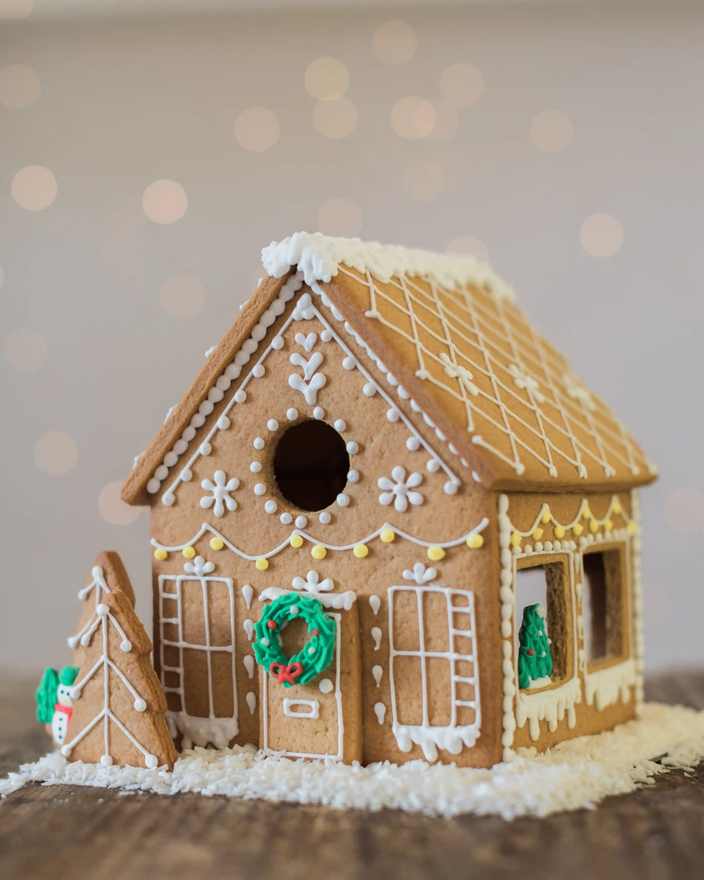 Honeywell Bakes Gingerbread House Baking Kit - A decorated gingerbread house with white icing details, a green wreath, and a small gingerbread tree beside it, set on a wooden surface with a soft blurred holiday background.