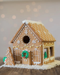 Honeywell Bakes Gingerbread House Baking Kit - A decorated gingerbread house with white icing details, a green wreath, and a small gingerbread tree beside it, set on a wooden surface with a soft blurred holiday background.