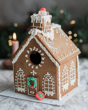Honeywell Bakes Gingerbread House Baking Kit - A decorated gingerbread house with white icing details, a green wreath on the door, and small red and green sugar decorations, photographed in a softly blurred holiday setting with lights and a candle in the background