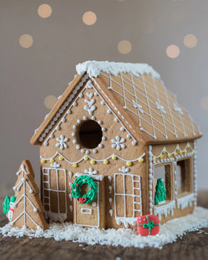 Honeywell Bakes Gingerbread House Baking Kit - A decorated gingerbread house with white icing details, a green wreath on the front door, a small gingerbread tree beside it, and red gift decorations on a snowy base, photographed against a softly blurred warm background.