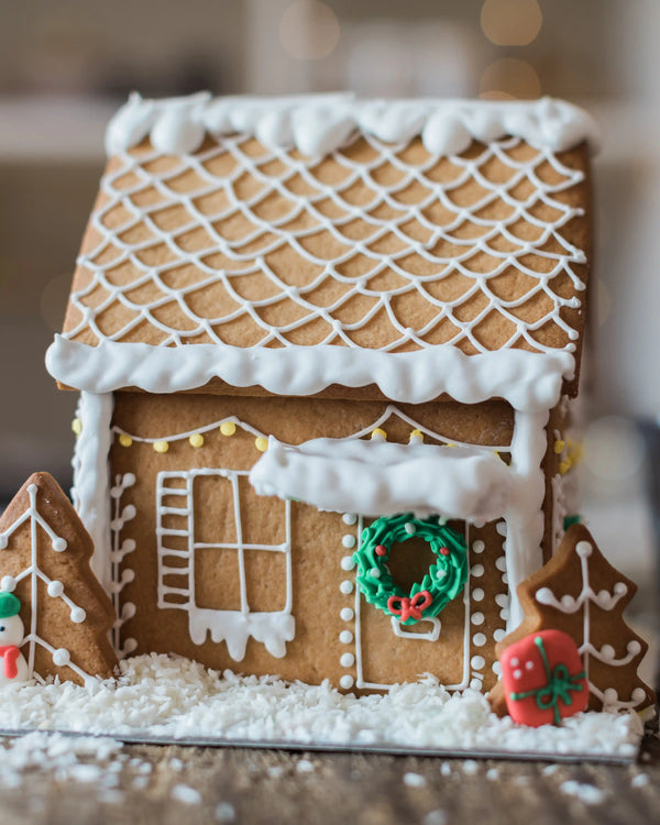 Honeywell Bakes Gingerbread House Baking Kit - A decorated gingerbread house with white icing snow, a green wreath on the door, and small Christmas tree decorations, photographed in a warm blurred indoor setting.