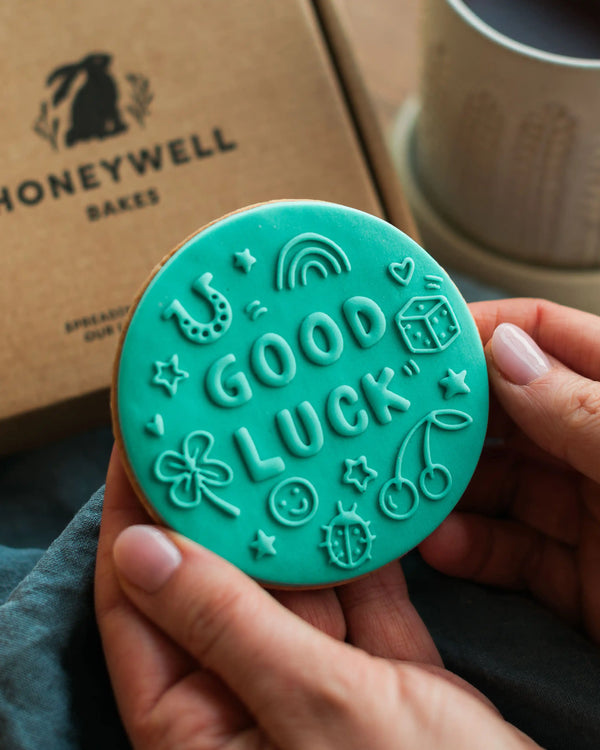 Honeywell Bakes Good Luck Biscuit - Teal cookie with 'Good Luck' text held by a person, with a Honeywell Bakes box in the background.
