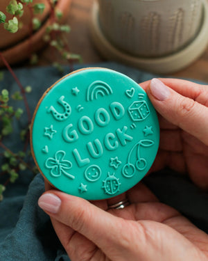 Honeywell Bakes Good Luck Biscuit - Teal round object with 'Good Luck' text held by a hand, with a blurred background