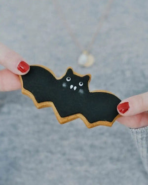 Honeywell Bakes Halloween Biscuit Gift Set - Bat-shaped cookie with black icing held between fingers against a blurred background