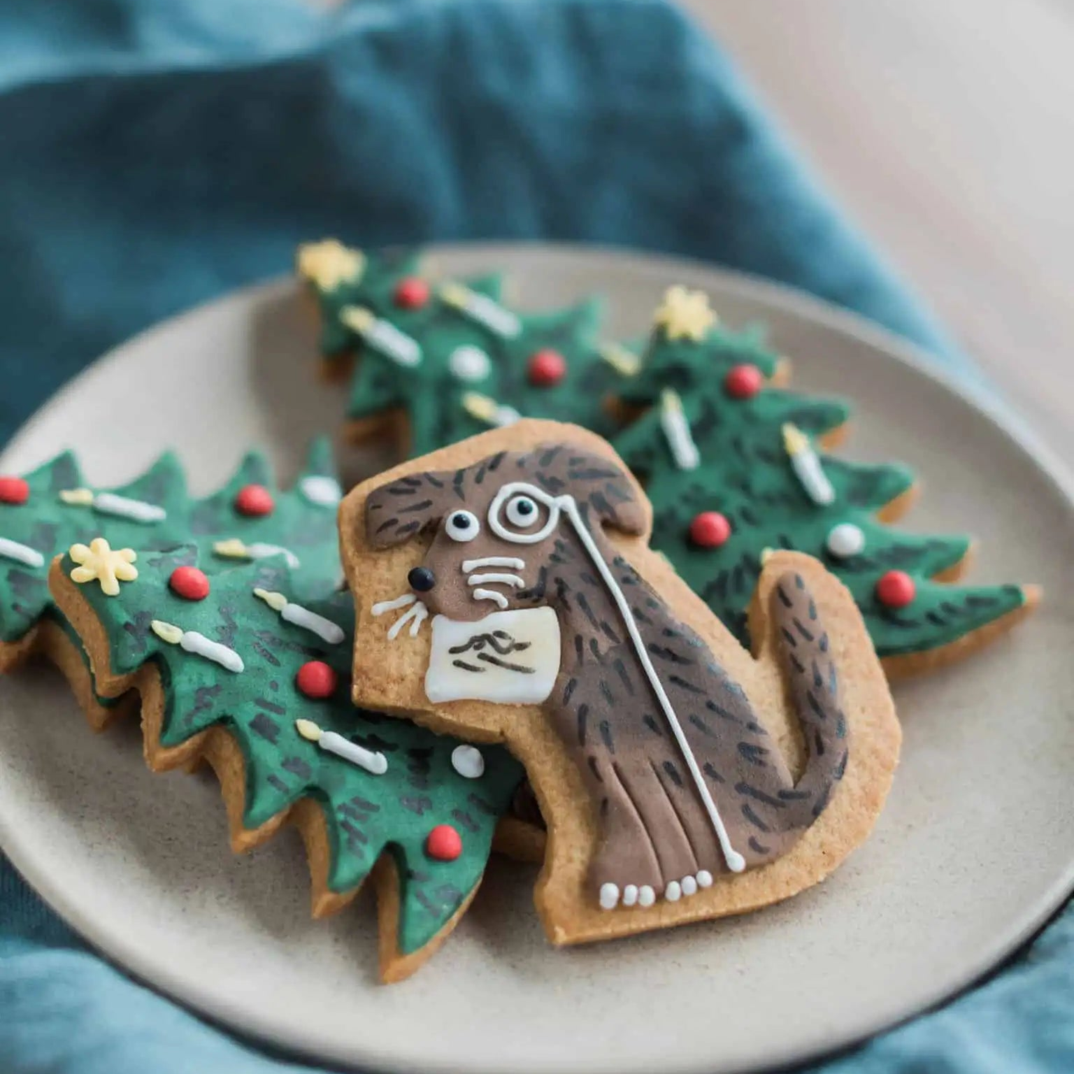 Decorative cookies shaped like a dog and Christmas trees on a plate with a blue cloth background.  Made for Axel Scheffler Children's Illustrator for Julia Donaldson books
