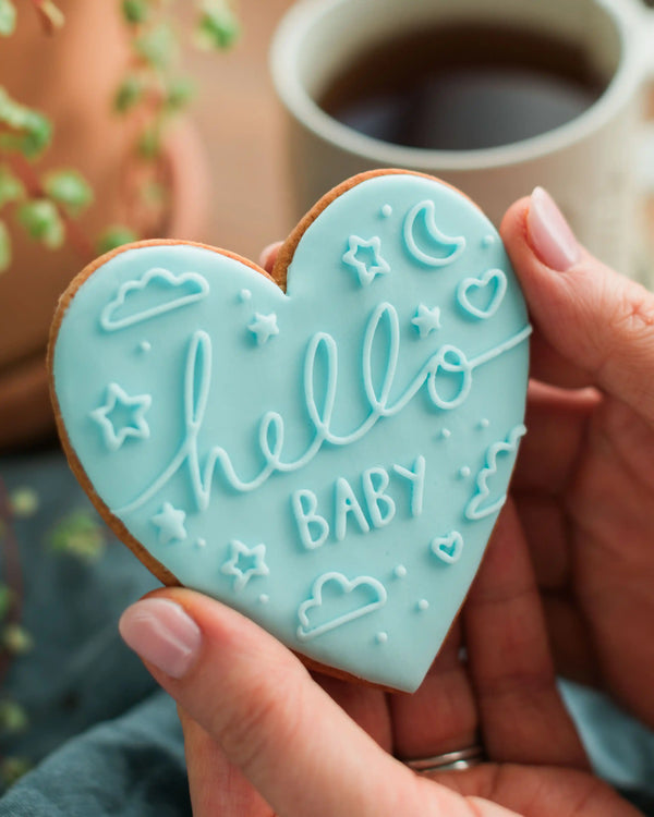 Honeywell Bakes Hello Baby Biscuit Blue - Heart-shaped cookie with 'hello baby' text held by a hand, with a blurred coffee cup in the background.