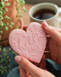 Honeywell Bakes Hello Baby Biscuit Pink - Heart-shaped cookie with 'hello baby' text held by a hand, with a cup of coffee and plant in the background.